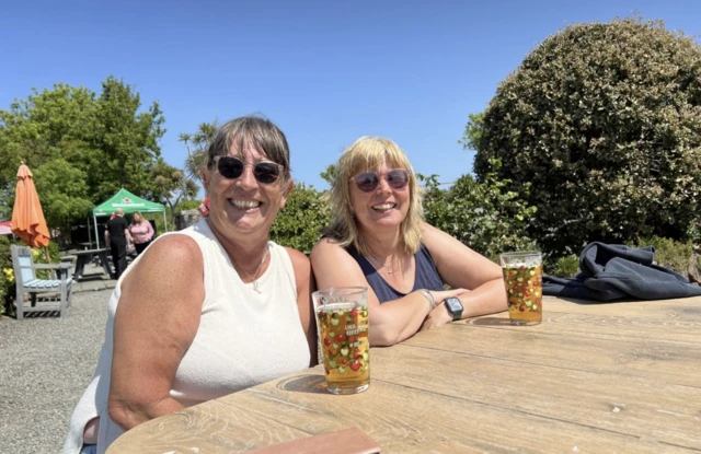 Karen Hollyhawk and Theresa Banks celebrating Liberation Day with a drink at the Portelet Inn, in Jersey. They are looking towards the camera smiling with drinks in front of them. There is a gazebo in the background. The sky is blue and there are trees behind the pair as well.