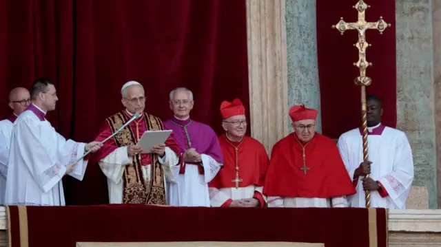 Pope Leo speaks from the balcony reading from a paper and speaking into a microphone. He is surrounded by men dressed in religious clothing.