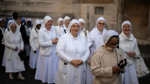 Group of white-clad nuns outside a stone church, smiling and waiting to attend mass.