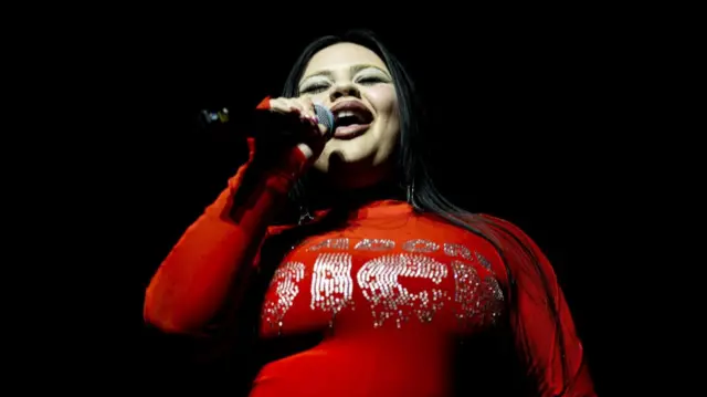 Miriana Conte performs in a long-sleeved red shirt with rhinestones on the front. She has her eyes closed and is holding a mic with her right hand raised towards her mouth