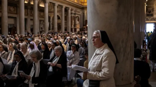 Nuns gathered in grand marble church, reading from prayer book. In foreground is nun in a white jacket standing in front of a pillar.