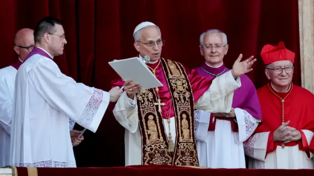 Pope Leo speaks into a microphone surrounded by other men in religious clothing. He speaks from the  balcony of St. Peter's Basilica with his arms widened.