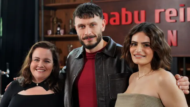 Jessica Gunning (L), Richard Gadd (C) and Nava Mau (R) standing together smiling. Behind them is a replica of a wooden bar counter with bottles on the shelves and a sign reading Baby Reindeer with the Netflix logo in red