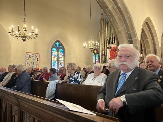 People are sat in a church service. There are a number of people sat down on benches with the church design found in the background.