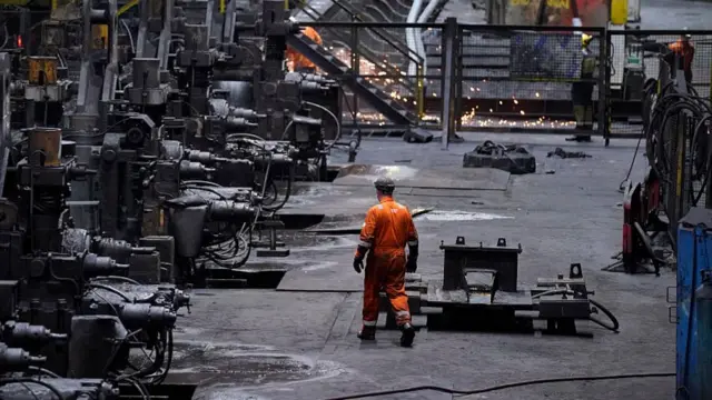 A steel worker in an orange jumpsuit, walking on factory floor surrounded by machinery.