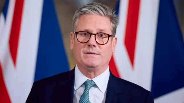 Starmer with UK flags behind him, staring into camera wearing blue suit, blue tie and glasses.