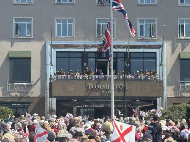 Re-enactors dressed as WW2 soldiers on the balcony of a hotel wave to the crowd - many of whom are waving flags