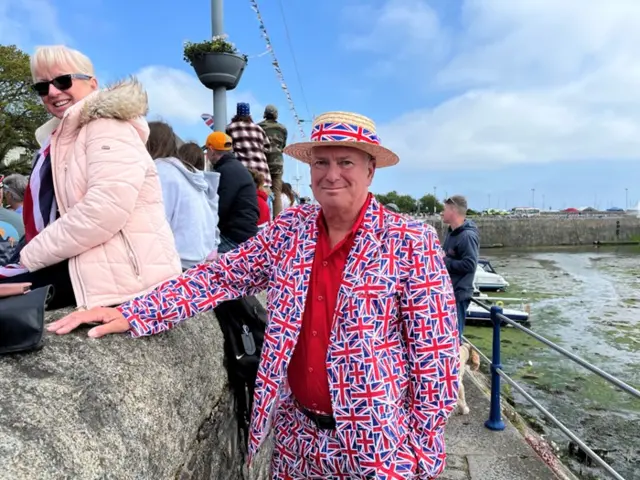 A man is wearing a suit covered in mini Union Jack flags. He is wearing an all red suit and a hat.