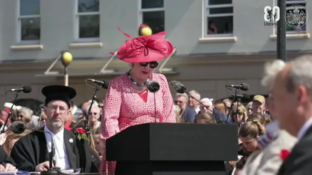 St Martin Constable Karen Shenton Stone giving a speech in Liberation Square in St Helier, Jersey. She is wearing a pink dress and a pink hat, while stood at a podium speaking to a crowd.