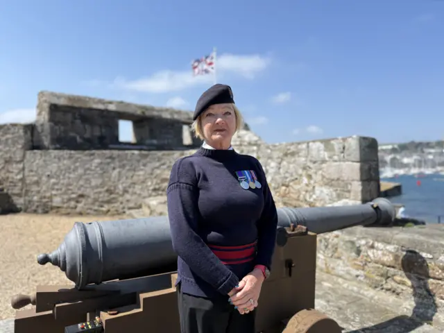 A woman in a blue woollen jumper with medals on and a blue beret stands in front of a cast iron canon on the battlement of a castle.