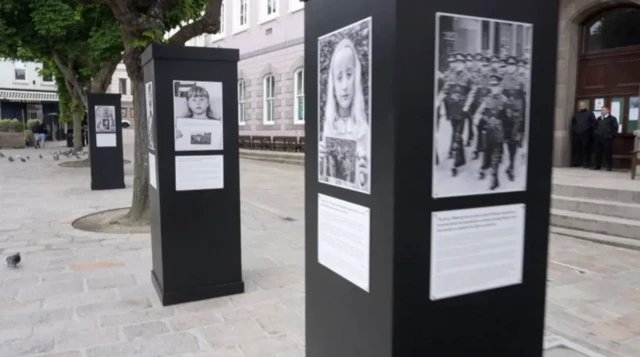 A picture of plinths in Jersey's royal square. They are black with black and white images on. They are surrounded by buildings and trees.