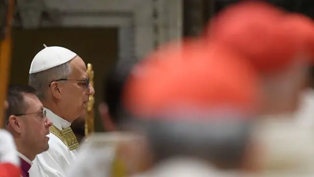 Pope with eyes closed, speaking, in background, whilst out-of-focus heads of cardinals wearing red caps in foreground.