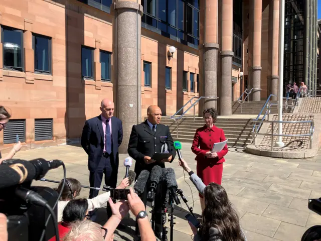 Supt Kevin Waring wearing his police uniform stands in between a man and a woman to read his statement to the media in front of Newcastle Crown Court. A number of reporters holding microphones are on the ground in front of him