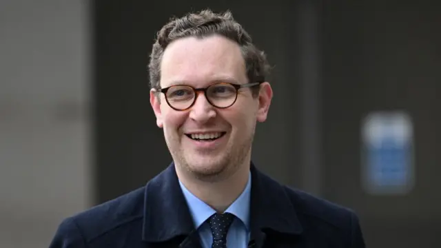 Darren Jones wearing suit and coat, smiling at camera. Blurred background.