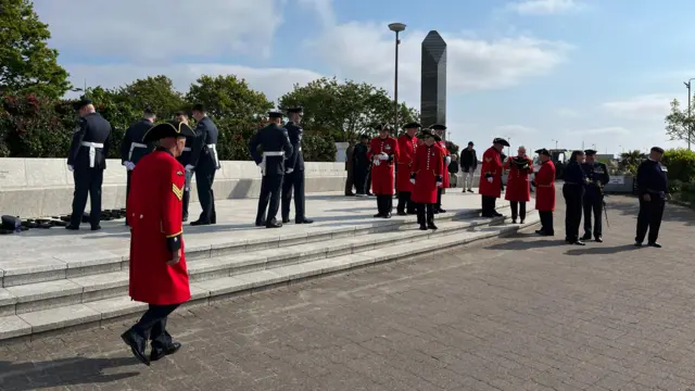 About two dozen men in ceremonial military uniforms gather near a tall concrete obelisk. They include Chelsea Pensioners in red jackets and tricorn hats.