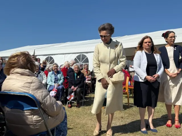 Princess Anne met people who stayed in the occupation during the war. She is wearing a cream colour outfit and glasses. People are sat and stood around her.