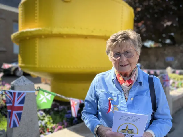 Pamela is wearing a blue outfit and glasses. She is stood in front of a yellow design and flowers with flag banners.