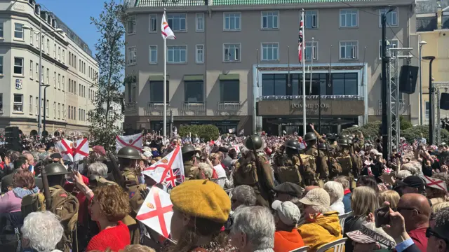 Re-enactors dressed as WW2 soldiers march through a crowd waving flags