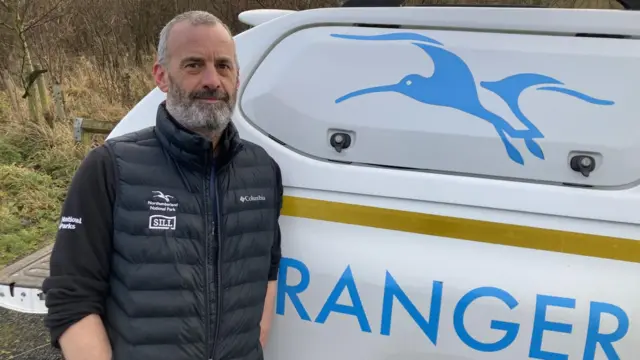Gary Pickles leaning against a car with the word ranger on it and a blue bird logo. He is wearing a black gilet with a Northumberland National Park logo and a black top with the sleeves rolled up and he has short grey hair and a beard