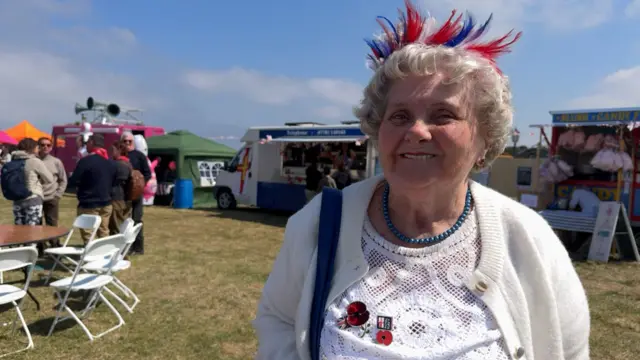 An white-haired lady wearing a cardigan in a field with a bright red, white and blue head dress on. Behind are food trucks.