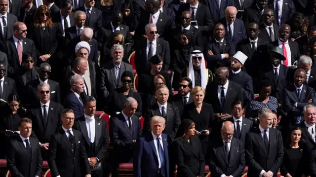 A number of world leaders can be seen standing and dressed in dark clothing. Donald Trump is among those in the front row with his wife Melania.