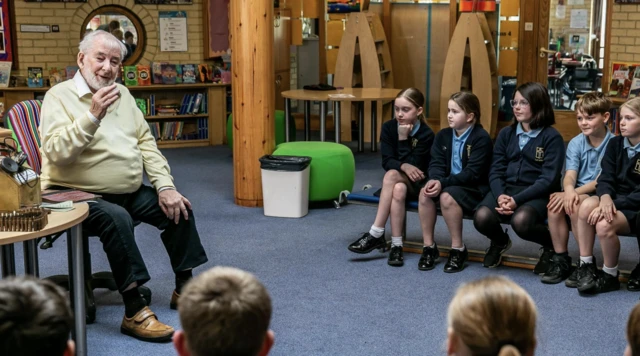 François Le Maistre sits on a chair as he talks to a group of school children sat in a semi-circle around him. Some World War Two artefacts are on the table alongside him.