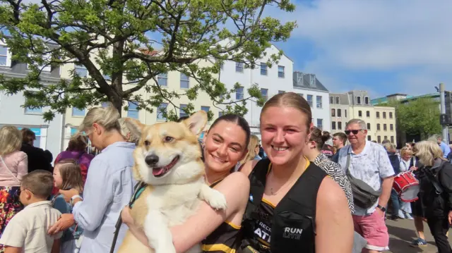Two smiling young women in black singlets. One has dark hair and is holding a large corgi, while the other has blonde hair.