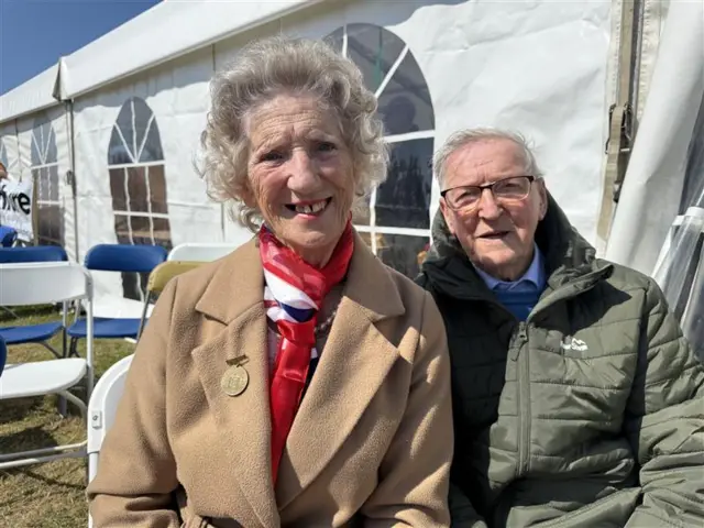 An old married couple in coats in front of a white marquee.