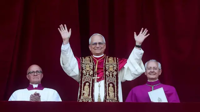 Pope Leo XIV stands in a balcony in ornate robes and spreads his arms in celebration