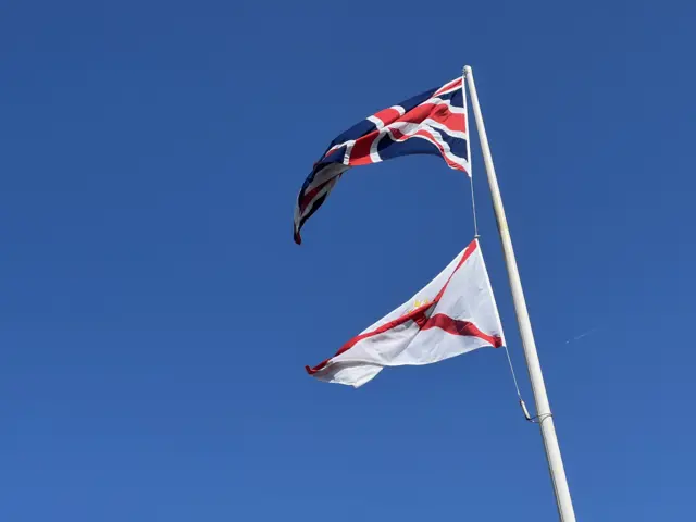 Two flags are raised on a white flagpole. The blue sky can be seen behind it.