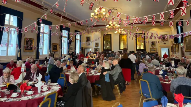 People sat in a room around tables. There are bunting strings with flags on hanging from the ceiling.