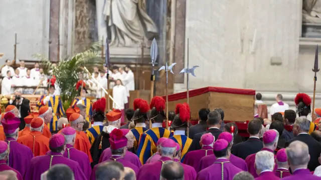 Pope Francis' coffin is being carried by members of the Swiss Guard towards the altar. Many men can be seen dressed in purple, red and white.