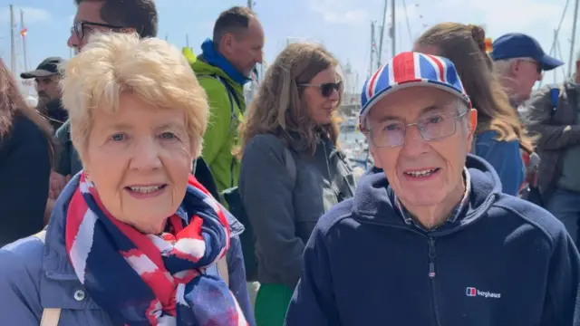 A married couple in their 80s wearing blue. The woman wears a Union Flag scarf while the man wears glasses and a Union Flag cap.