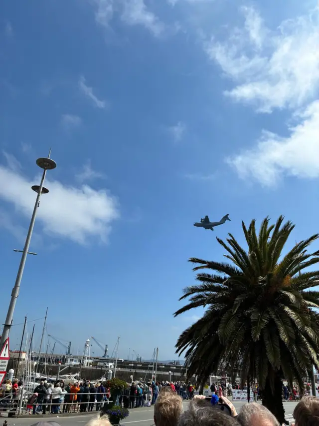 A military plane flies above a harbour and palm tree.