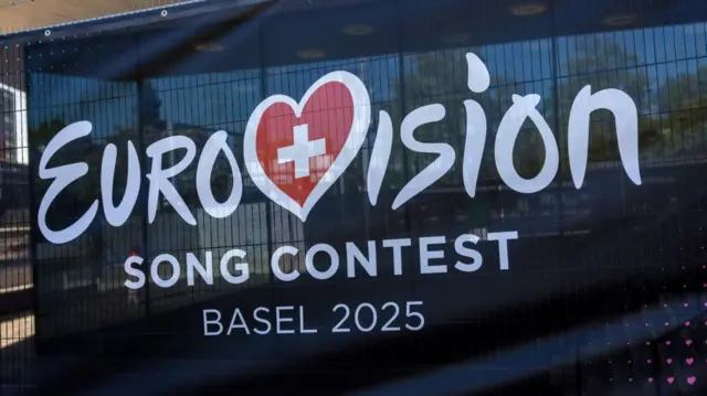 Dark blue festoon held up by metal barriers around St Jakobshalle in Basel. On the festoon is the Eurovision logo, the V taking the shape of a heart with inside the Swiss flag. Underneath the wording Song Contest Basel 2025 in white print