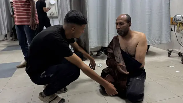 A man with his chest exposed sits on the floor and is being spoken to by another person