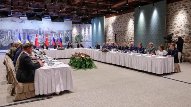 Ukrainian and Russian delegations meet in Turkey for talks. they sit at long white tables. about 20 people are shown, men and women, in a room with stone walls. ukrainian, russian and turkish flags can be seen.