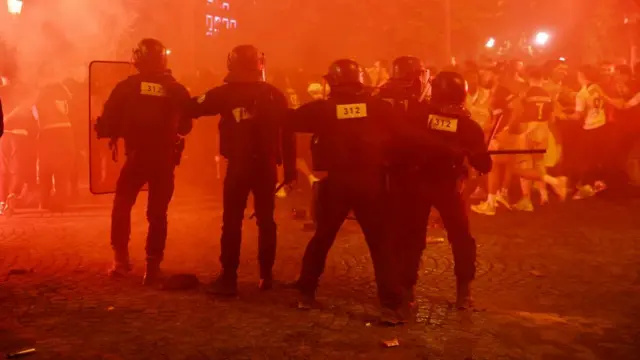Paris riot police stand facing a crowd of people, with smoke and red light illuminating them