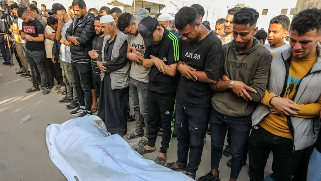 Men bow their heads standing beside a white body bag on the ground