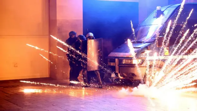 Police hide behind a riot shield as a firework sparks in front of them