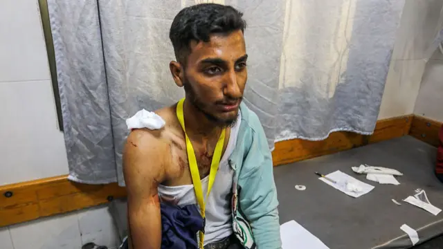 A man sits on a hospital bed, with torn clothing and blood stains on his chest
