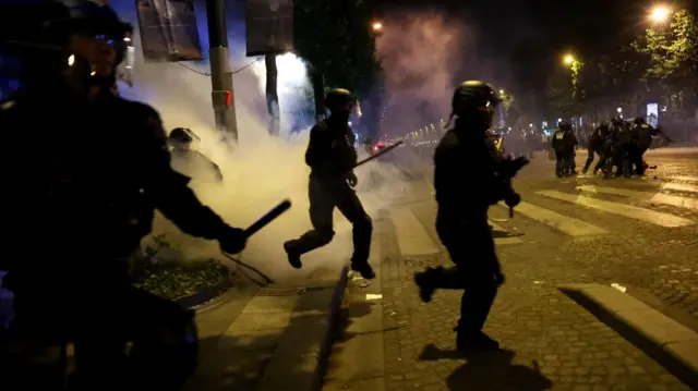 French police run into a road with truncheons drawn and smoke behind them