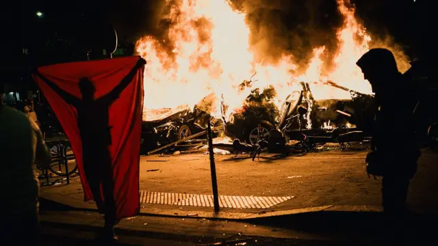 Person holds a flag behind them as they stand in front of cars on fire