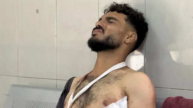 A man with a beard grimaces, topless with bandages in his torso