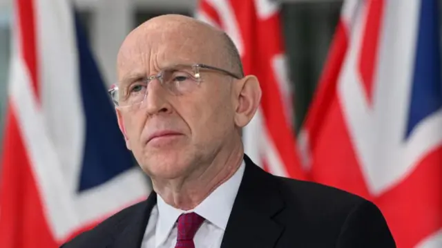 Defence Secretary John Healey wears a suit and red tie with UK flags in the background.