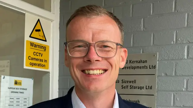 Ashley Shorey-Mills smiles at the camera while standing in a building doorway. He has short blonde hair and wears glasses with a navy suit jacket and a white shirt.