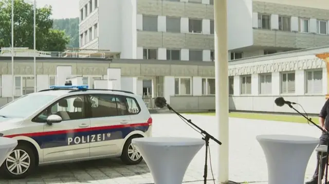 A still from outside a school where there are empty podiums with microphones in front. The polizei car is seen behind