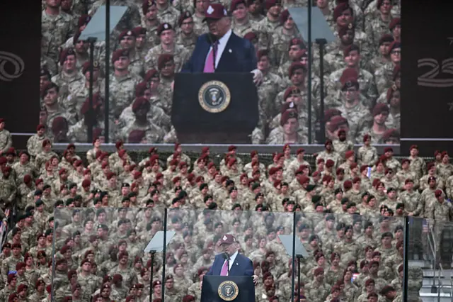 Donald Trump is seen speaking in front of a large military rally, and a large screen behind him also shows him speaking in the picture. He wears a maroon coloured hat - different from his traditional red Maga cap - that seems to match the red berets worn by the military personnel in the crowd.