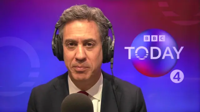 Ed Miliband is wearing headphones and sitting at a microphone. he is wearing a black suit. behind him is a red and blue background with the bbc logo for today