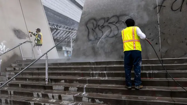 Two men wearing high-vis jackets use a hose to remove graffiti from grey walls